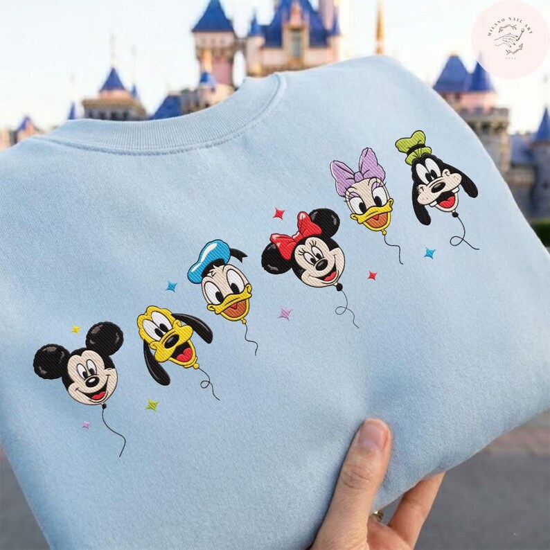 May include: Light blue sweatshirt featuring embroidered Disney characters as balloons. Mickey Mouse, Pluto, Donald Duck, Minnie Mouse, Daisy Duck, and Goofy are depicted as colorful balloons with star accents. A fun and whimsical design.