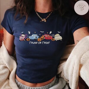 May include: A navy blue short-sleeve t-shirt featuring a graphic of four cartoon cars dressed as Halloween characters, including a vampire, a mummy, and a red race car, with orange pumpkins, white ghosts, and the text TRUNK OR TREAT.