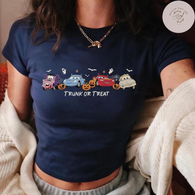 May include: A navy blue short-sleeve t-shirt featuring a graphic of four cartoon cars dressed as Halloween characters, including a vampire, a mummy, and a red race car, with orange pumpkins, white ghosts, and the text TRUNK OR TREAT.
