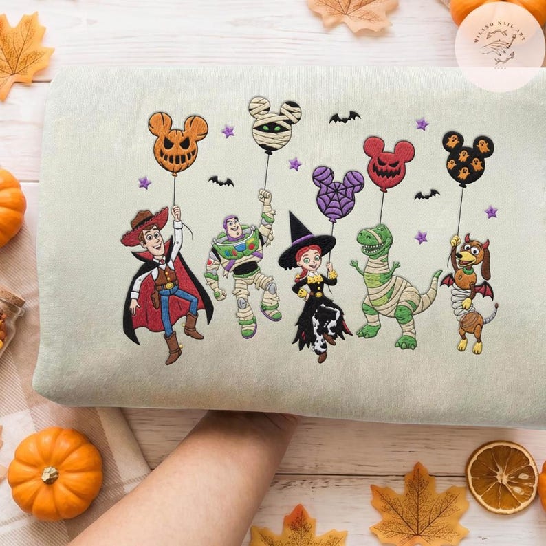 May include: A light green sweatshirt featuring embroidered Toy Story characters in Halloween costumes: Woody as a vampire, Buzz Lightyear as a mummy, Jessie as a witch, Rex as a mummy, and Slinky Dog as a devil, all holding Mickey-shaped balloons.