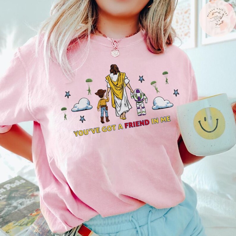 May include: Pale pink crew neck t-shirt with a graphic of Jesus, Woody, and Buzz Lightyear. The design includes clouds, stars, and parachutes, with the text "YOU'VE GOT A FRIEND IN ME". A white mug with a smiley face is held in the hand.