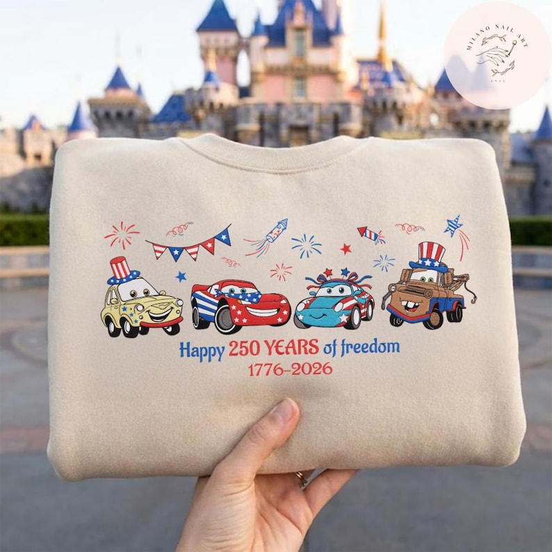 Pode incluir: Camisola bege com carros de desenho animado com designs patri&oacute;ticos e fogos de artif&iacute;cio. O texto "Happy 250 Years of freedom 1776-2026" est&aacute; impresso abaixo dos carros. A camisola &eacute; segurada por uma pessoa.