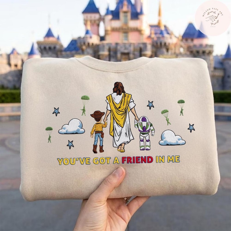 May include: Beige sweatshirt with a graphic of a figure in a white robe and yellow shawl, holding hands with a cartoon cowboy and Buzz Lightyear. The text "YOU'VE GOT A FRIEND IN ME" is printed below the figures. Stars, clouds, and parachuters are also featured.