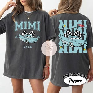 May include: Dark grey t-shirt with a light blue graphic featuring the word "MIMI", a cartoon car, and the word "CARS". The back of the shirt repeats the word "MIMI" in a pattern with a car graphic.