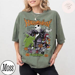 May include: Olive green t-shirt featuring a Halloween-themed graphic with Jack Skellington and other characters. The design includes the text "This is Halloween" and "California Adventure". The shirt has short sleeves and a crew neck.