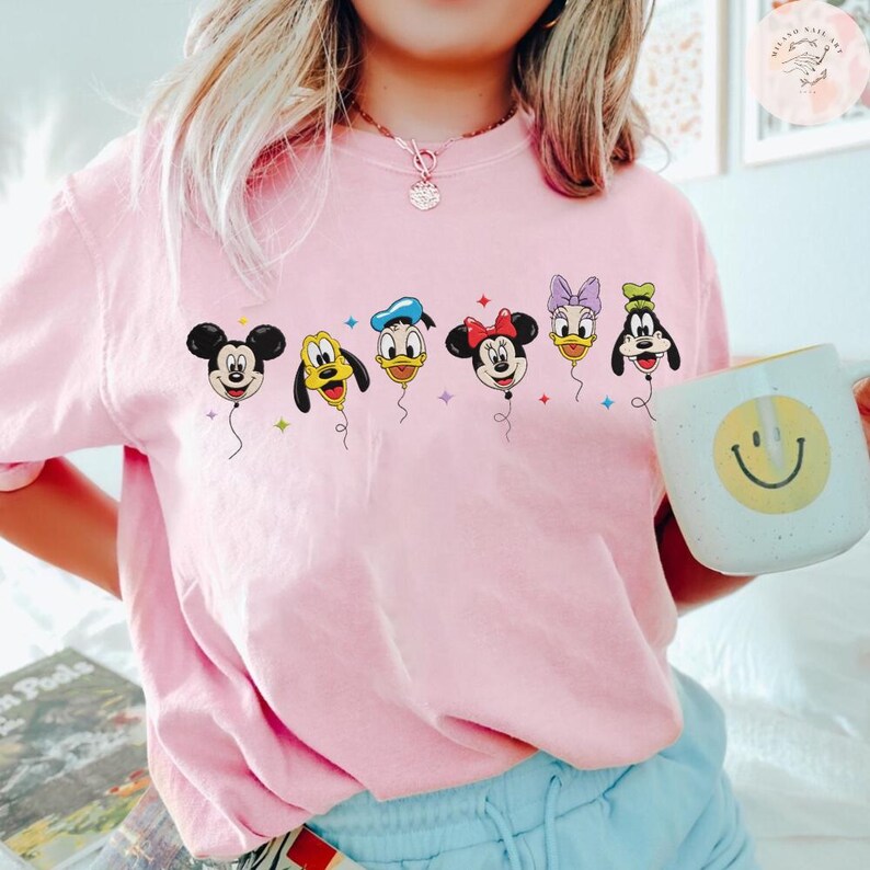 May include: A light pink sweatshirt featuring a colorful graphic of Disney characters as balloons. The balloons include Mickey Mouse, Pluto, Donald Duck, Minnie Mouse, Daisy Duck, and Goofy. A white mug with a smiley face is held in the hand.