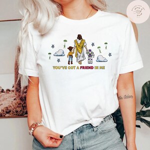 May include: White t-shirt with a graphic of Jesus, a cowboy, and a space ranger. The design includes clouds, stars, and the text "YOU'VE GOT A FRIEND IN ME" in yellow. A casual wear item.