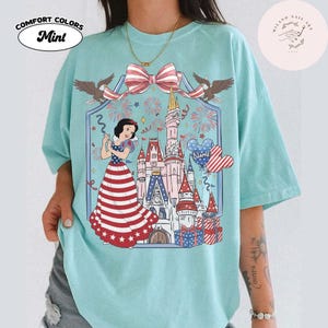 May include: Mint-coloured Comfort Colors t-shirt with a graphic of Snow White in a patriotic dress, a castle, fireworks, and a bow. The design includes two eagles and a heart-shaped balloon. The text "NAME" is on the balloon.