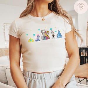 May include: Cream-colored t-shirt featuring a colorful graphic of characters from a popular animated movie. The design includes a crown, a dragon-like creature, two sisters, a snowman, and a castle. The shirt has short sleeves and a crew neck.