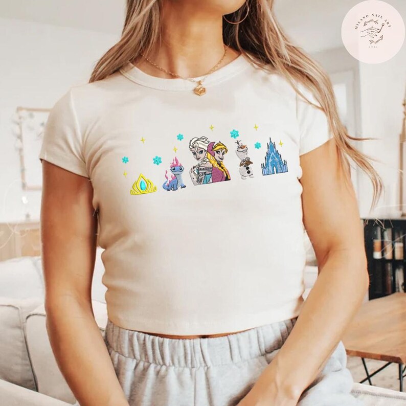 May include: Cream-colored t-shirt featuring a colorful graphic of characters from a popular animated movie. The design includes a crown, a dragon-like creature, two sisters, a snowman, and a castle. The shirt has short sleeves and a crew neck.
