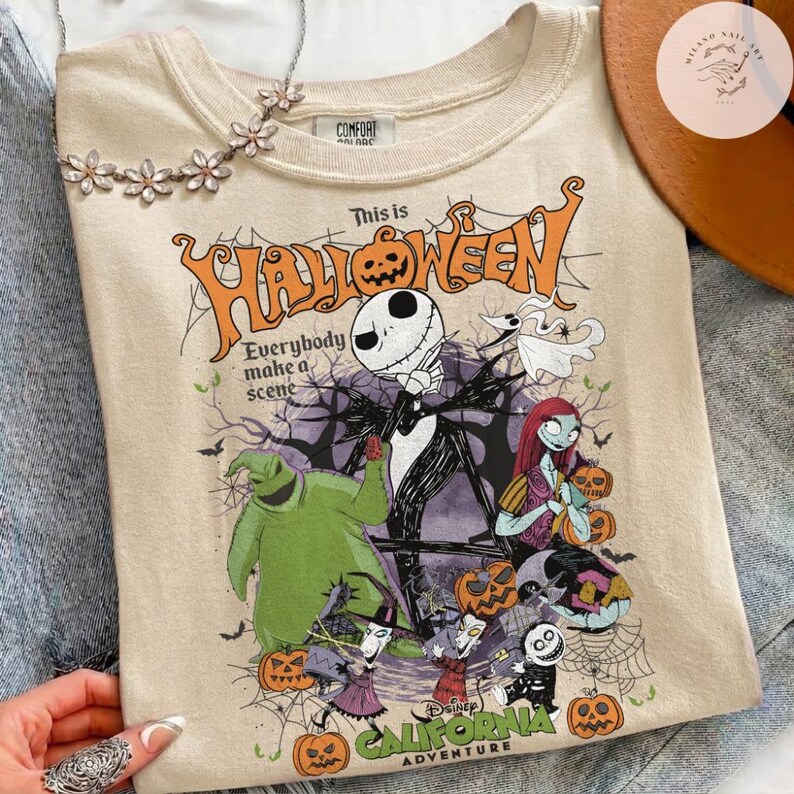 May include: Beige t-shirt featuring a colorful graphic from "The Nightmare Before Christmas." The design includes Jack Skellington, Sally, and other characters, with the text "This is Halloween" and "California Adventure."