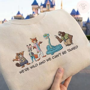 May include: Beige sweatshirt featuring embroidered characters from a popular animated film. The design includes a beaver, a rabbit, a fox, a snake, and a sloth. The text "WE'RE WILD AND WE CAN'T BE TAMED" is embroidered below the characters.