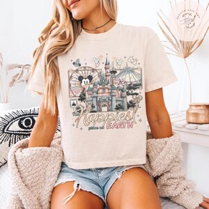 May include: Cream-colored t-shirt featuring a colorful graphic of a castle, a Ferris wheel, and other theme park elements. The text "The Happiest Place on Earth" is displayed in a script font. The shirt is a relaxed fit.