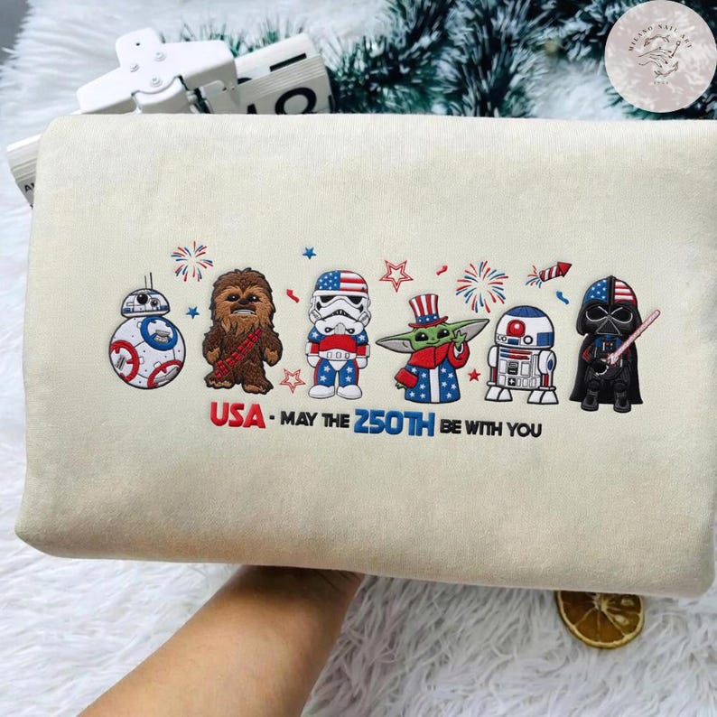 Op de afbeelding: Een cr&egrave;mekleurig kussen met geborduurde Star Wars-personages in patriottische kleding. Het ontwerp bevat BB-8, Chewbacca, een Stormtrooper, Grogu, R2-D2 en Darth Vader, met de tekst "USA - May the 250th be with you."