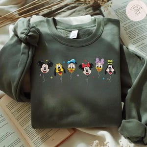 May include: An olive green sweatshirt featuring a colorful graphic of Disney characters as balloons. Mickey Mouse, Goofy, Donald Duck, Minnie Mouse, Daisy Duck, and Pluto are depicted. The sweatshirt is made of a soft material and has a crew neck.