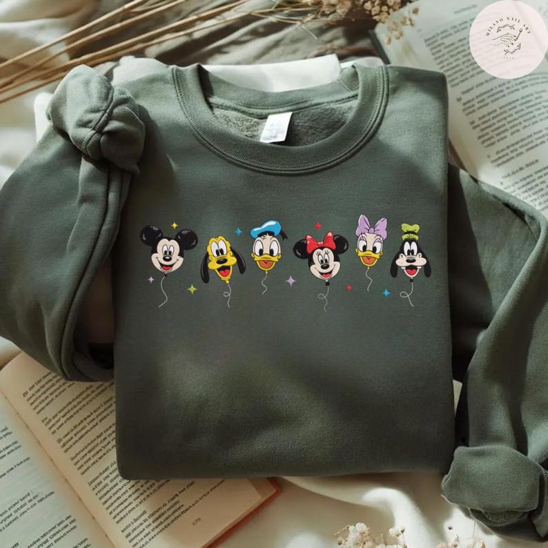 May include: An olive green sweatshirt featuring a colorful graphic of Disney characters as balloons. Mickey Mouse, Goofy, Donald Duck, Minnie Mouse, Daisy Duck, and Pluto are depicted. The sweatshirt is made of a soft material and has a crew neck.
