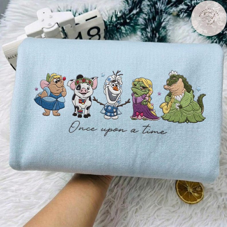 May include: A light blue sweatshirt featuring embroidered cartoon characters. The design includes a fairy, a pig, Olaf the snowman, a frog, and an alligator. The phrase "Once upon a time" is written in script below the characters. The sweatshirt is displayed on a white, fluffy surface.