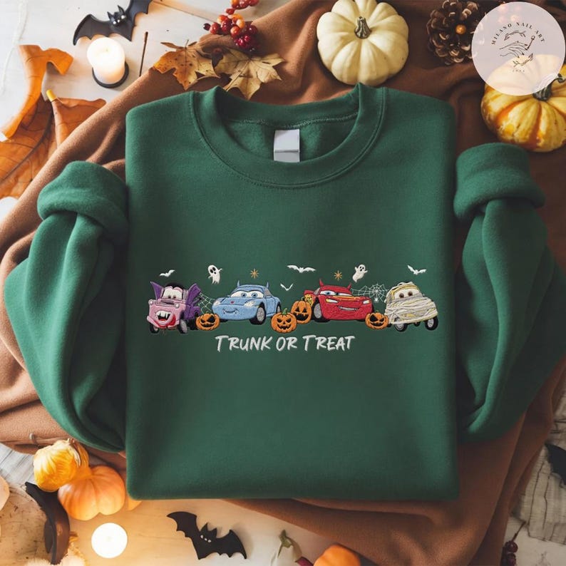 May include: A dark green crewneck sweatshirt featuring a graphic of four cartoon cars dressed in Halloween costumes, including a vampire, a mummy, and others, with orange jack-o'-lanterns and the white text "TRUNK OR TREAT" printed below.
