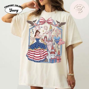 May include: Ivory Comfort Colors t-shirt with a patriotic design. The graphic features a princess in a red, white, and blue dress, a castle, fireworks, and a bow. The shirt has short sleeves.