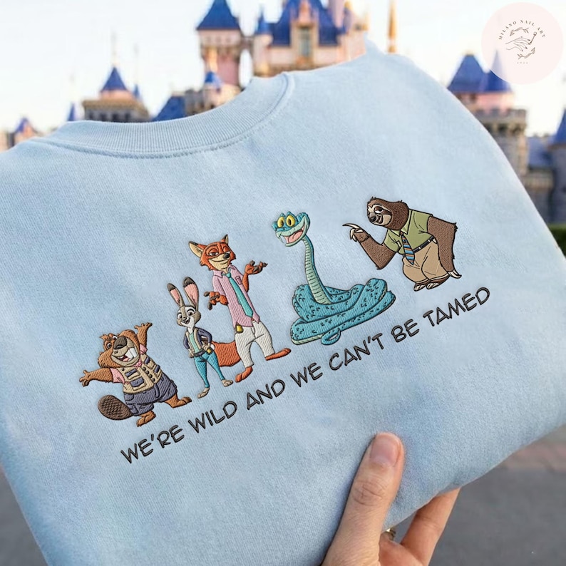May include: Light blue sweatshirt featuring embroidered cartoon characters from a popular animated film. The design includes a beaver, rabbit, fox, snake, and sloth. The text "WE'RE WILD AND WE CAN'T BE TAMED" is also embroidered on the sweatshirt.