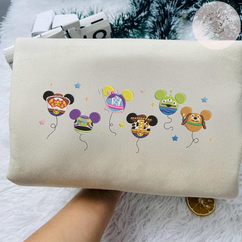 May include: A beige sweatshirt features colorful embroidered designs of cartoon character-themed balloons. The balloons are in the shape of Mickey Mouse heads, with various character-inspired designs and colors. Small star and plus-shaped embroidery accents are scattered around the balloons.