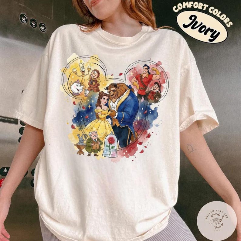 May include: An ivory t-shirt featuring a graphic print of characters from Beauty and the Beast arranged within a Mickey Mouse ear silhouette. The design includes Belle, the Beast, Gaston, and supporting characters, with a red rose in a glass dome.