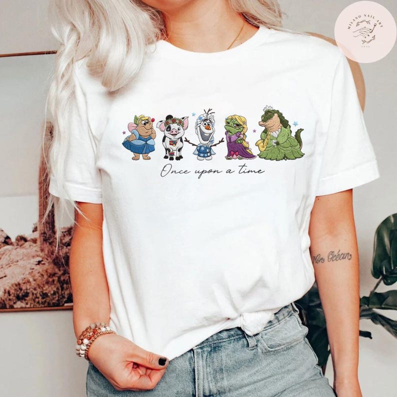 May include: White t-shirt featuring a whimsical illustration of cartoon characters. The design includes a fairy, a pig, Olaf, a princess, and a dragon. The phrase "Once upon a time" is written below the characters in a cursive font.