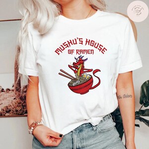 Op de afbeelding: Wit T-shirt met een cartoon draak die eetstokjes boven een kom ramen vasthoudt. De tekst "Mushu's House of Ramen" staat in het rood boven de afbeelding. Een casual, alledaags kledingstuk.