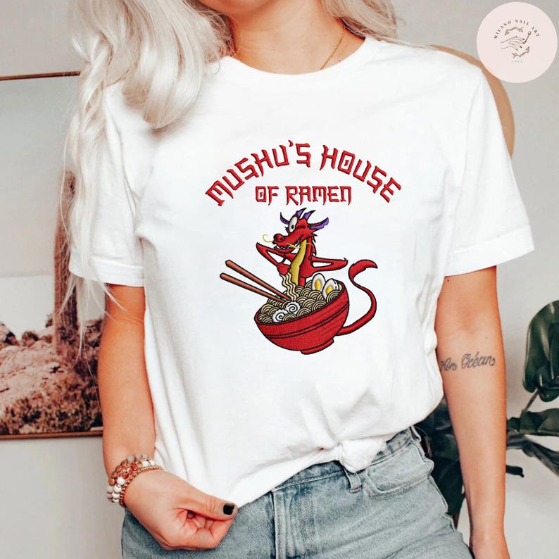 Op de afbeelding: Wit T-shirt met een cartoon draak die eetstokjes boven een kom ramen vasthoudt. De tekst "Mushu's House of Ramen" staat in het rood boven de afbeelding. Een casual, alledaags kledingstuk.