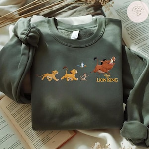 May include: An olive green sweatshirt with an embroidered design of characters from Disney's The Lion King. The design features Simba, Timon, Pumbaa, and Zazu. The words "The Lion King" are printed in gold. The sweatshirt is displayed on a neutral surface.
