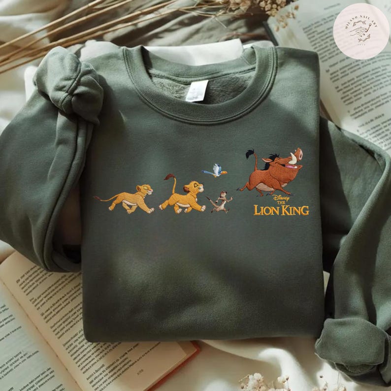 May include: An olive green sweatshirt with an embroidered design of characters from Disney's The Lion King. The design features Simba, Timon, Pumbaa, and Zazu. The words "The Lion King" are printed in gold. The sweatshirt is displayed on a neutral surface.