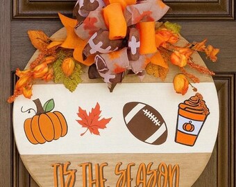 May include: A round wooden fall-themed door hanger with a white and light brown design. It features a pumpkin, maple leaf, football, and coffee cup graphic. The text "Tis the Season" is displayed in orange. The top is decorated with orange and brown ribbons and fall foliage.