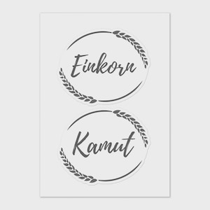 May include: Two circular stickers with the words "Einkorn" and "Kamut" in a handwritten font. Each sticker has a wheat stalk design around the edge, and the background is a light grey.