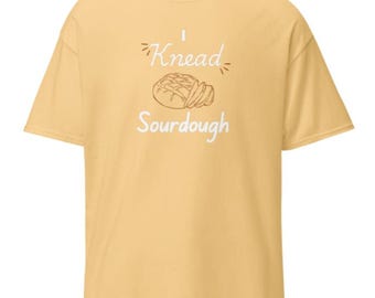 May include: A light yellow t-shirt with the text "I Knead Sourdough" in white. The design includes a line drawing of a loaf of bread. The shirt has short sleeves and a crew neck.