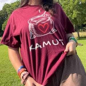 May include: A maroon t-shirt featuring a white graphic of a container with a heart and the word "KAMUT". The shirt is being held up, revealing the lower portion of the person's body. The background is a grassy area with trees.