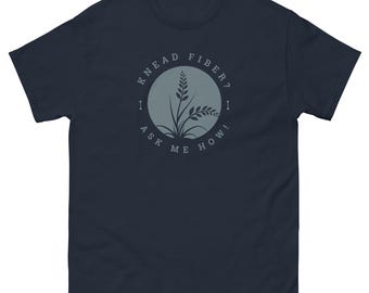 May include: Navy blue t-shirt with a circular graphic featuring a plant design and the text "KNEAD FIBER? I ASK ME HOW!". The design is in a muted gray color. The shirt is made of a soft, comfortable material.