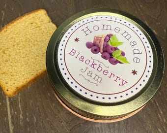 May include: A jar of homemade blackberry jam with a gold lid and a label featuring a blackberry illustration. A slice of golden bread sits next to the jar on a wooden surface.