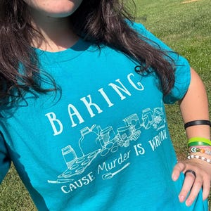 May include: Teal t-shirt with the words "BAKING CAUSE MURDER IS WRONG" in white. The shirt features a graphic of baking supplies. The shirt is short-sleeved.
