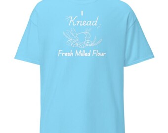 May include: A light blue t-shirt with the word "Knead" in white script above a graphic of wheat and bread. Below the graphic, the text "Fresh Milled Flour" is printed in white. The shirt has short sleeves and a crew neck.