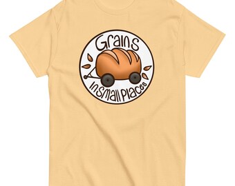 May include: A light yellow t-shirt featuring a circular graphic with the words "Grains in small places." The graphic depicts a loaf of bread on a small cart with wheels, surrounded by leaves. The shirt is made of a soft material.