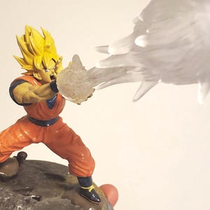 May include: A yellow-haired anime character figurine in an orange jumpsuit, with a blue belt, is firing a white energy blast from their hand. The figurine is standing on a rocky base.