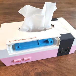 May include: A white and pink tissue box shaped like a retro video game console. The box has a blue slot for tissues and says "ENTERTAINMENT SYSTEM" on the front. It has buttons labelled "POWER" and "RESET".