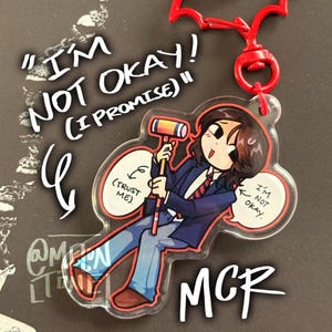 May include: A clear acrylic keychain with a cartoon illustration of a person holding a gavel. The keychain has a red bat-shaped clip and the text "I'M NOT OKAY! (I PROMISE)" and "MCR". The character wears a blue blazer and jeans.