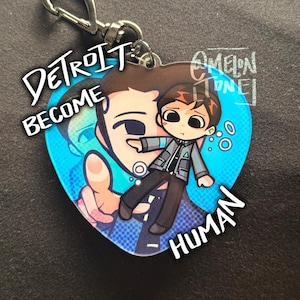 May include: Heart-shaped keychain featuring a cartoon illustration of a character from "Detroit: Become Human." The design includes the text "Detroit Become Human" and a character in a gray jacket. The background is blue with white bubbles.