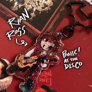 Puede incluir: Un llavero acrílico transparente con una ilustración de dibujos animados de un músico tocando la guitarra. El llavero tiene un cierre negro y el texto "Panic! At The Disco" y "Ryan Ross" son visibles. El fondo es rojo.