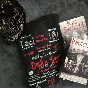 May include: A black sweatshirt with white text that reads "Devil's Night" and "Hosted by Four Horsemen". The sweatshirt is lying on a grey surface with a black mask and two books. The books are titled "Kill Switch" and "Nightfall" by Penelope Douglas.