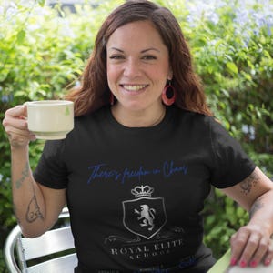Puede incluir: Una camiseta negra con un gráfico plateado de un león en un escudo con el texto "There's freedom in Chaos" y "Royal Elite School" debajo.