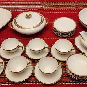 May include: A white porcelain tea set with gold trim. The set includes a teapot, creamer, sugar bowl, six teacups, six saucers, and a serving platter. The set is arranged on a red and white striped tablecloth.