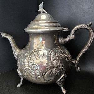 May include: A silver teapot with an ornate design and a bird finial on the lid. The teapot has three legs and a curved handle.