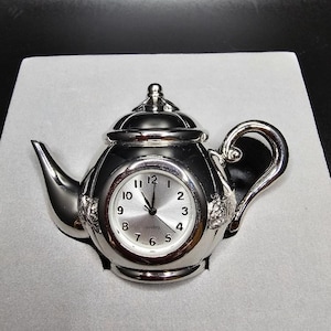 May include: A silver teapot-shaped clock with a white face and black hands. The clock reads "Quartz" and has black Roman numeral markings.